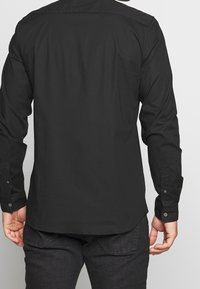 Czarna koszula z długim rękawem o gładkiej fakturze, z guzikami na mankietach i zaokrąglonym dołem, widziana z tyłu.