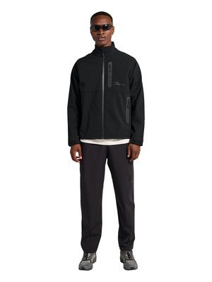 Man draagt een zwarte jas met ritssluiting en twee verticale zakken met rits, zwarte broek, grijze sneakers en zwarte zonnebril, staand met het gezicht naar voren.