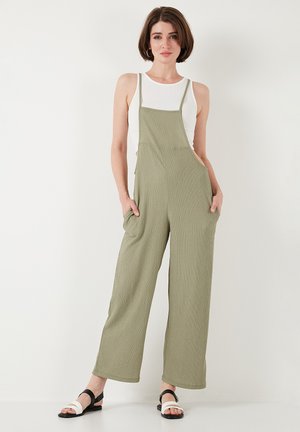 Roheline ribaga jumpsuit laiade jalgade ja küljetaskutega, millel on reguleeritavad rihmad ja lõdvestunud istuvus. Kombineeritud mustade ja valgete sandaalidega.