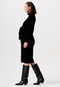 Zwarte sweaterjurk met hoge kraag, tailleband en lange mouwen; gecombineerd met kniehoge zwarte leren laarzen. Glad textuur, getailleerd silhouet.