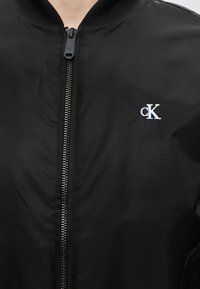 Veste noire zippée avec col côtelé et logo "cK" blanc brodé sur la poitrine gauche.