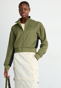 Oliven grønn zip-up jakke med svarte detaljer, høy krage og logo; kombinert med en kremfarget skjørt med lommer og elastisk band.