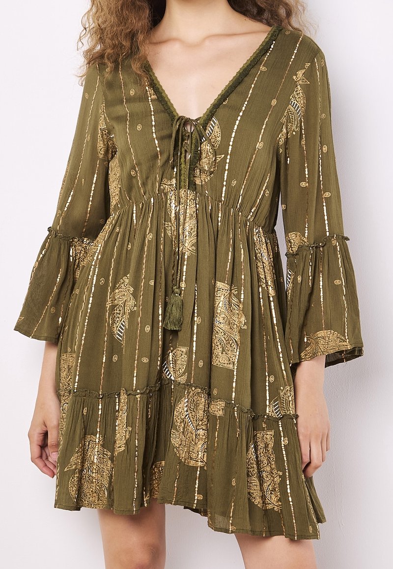 Femme portant une robe vert olive avec des motifs cachemire dorés et des rayures verticales, manches cloche, décolleté en V noué avec des cordons à pompons, et taille froncée.