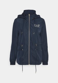 Mörkblå vindjacka med hög krage, dragsko i midjan och dragkedja. Har guldfärgad metalldetalj och logotyp på bröstet.