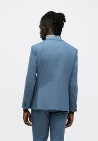 Világoskék zakó sima textúrával, félköríves hajtókával és két gombbal; megegyező színű nadrággal és lazán, nyitott szabással párosítva.