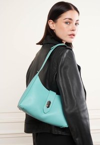Borsa a mano in pelle teal con una finitura testurizzata, caratterizzata da una forma triangolare e una chiusura in metallo argentato, indossata sopra una giacca di pelle nera.