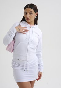 Felpa bianca con zip e coulisse, abbinata a una mini gonna bianca arricciata. Accessoriata con una piccola borsa rosa. Tessuto di texture liscia.
