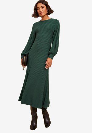 Grünes, geripptes Maxikleid mit langen Puffärmeln, tailliertem Schnitt und einem fließenden Rock. Kombiniert mit knielangen braunen Stiefeln mit Absatz und einer kleinen schwarzen Clutch.