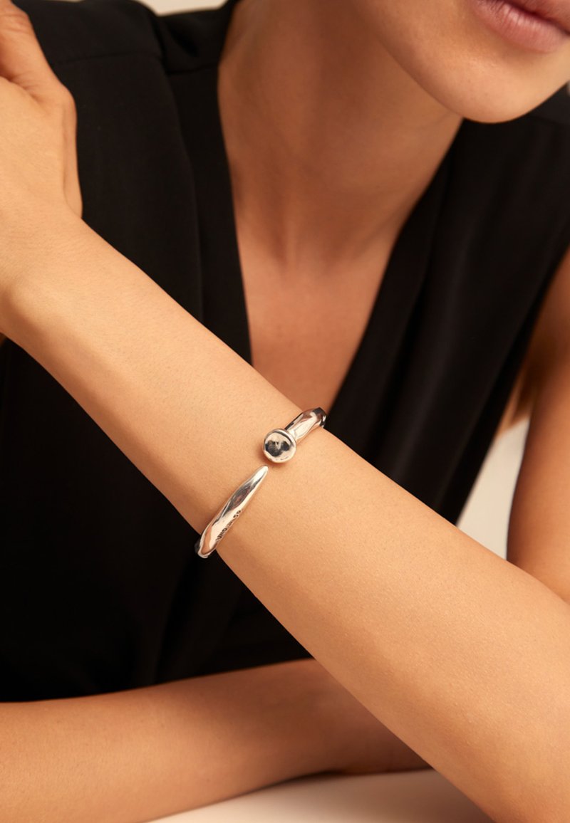 Bracelet manchette en métal argenté au design lisse et incurvé, orné d'un accent sphérique et d'une extrémité effilée. Surface texturée visible.