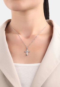 Ciondolo a croce d'argento su una catena sottile indossato intorno al collo sopra una maglietta bianca e il colletto di un blazer beige, con pelle e linea della mascella visibili.