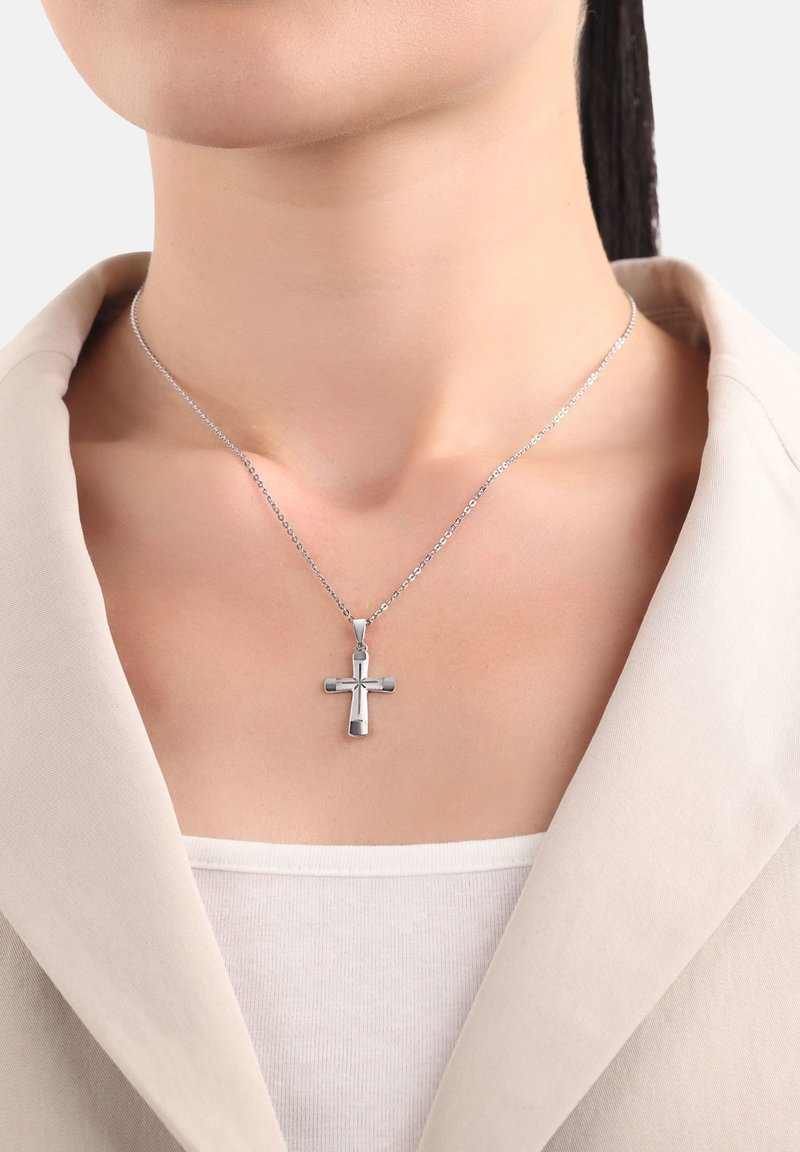 Ciondolo a croce d'argento su una catena sottile indossato intorno al collo sopra una maglietta bianca e il colletto di un blazer beige, con pelle e linea della mascella visibili.
