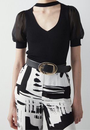 Top negro acanalado con mangas abullonadas transparentes y escote en V, combinado con pantalones de tiro alto con patrones abstractos en blanco y negro y cinturón con hebilla dorada.