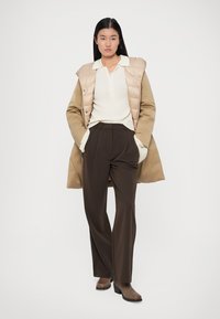 Beige puffer vest over en ribbet creme top, parret med højtaljede brune bukser. Modellen har brune ankelstøvler på og hænderne i lommerne.