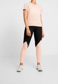 Světle růžové atletické tričko s mesh rameny, doplněné černými a světle růžovými legínami s geometrickým výřezovým designem.