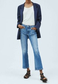 Blazer navy, camiseta blanca, jeans acampanados de mezclilla desgastada en azul y sandalias negras con tiras. La mezclilla tiene un dobladillo deshilachado, con un aspecto general casual.