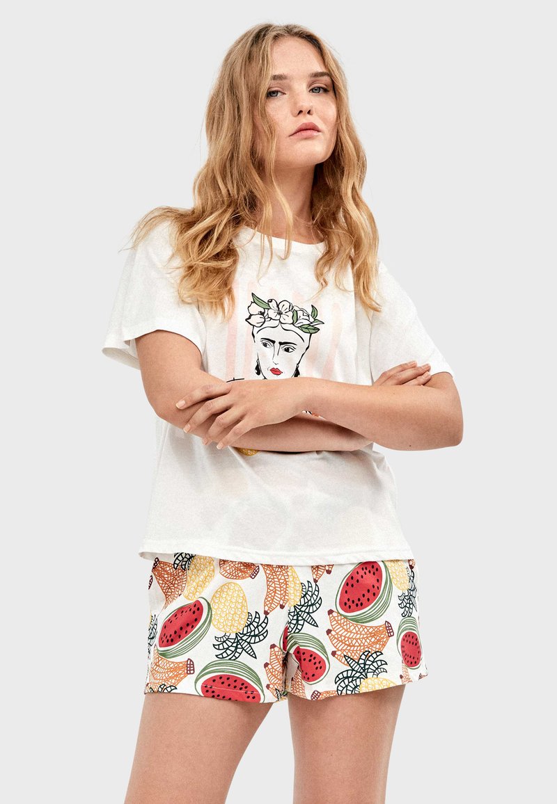 Jeune femme aux longs cheveux blonds portant un t-shirt blanc avec une illustration de visage et un short coloré à motifs de fruits, les bras croisés.