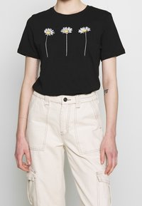 Camiseta de algodón negra con tres impresiones gráficas de margaritas blancas. Combinada con pantalones cargo beige claros con detalles de costura visibles.