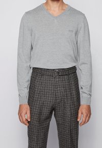 Grauer V-Ausschnitt-Pullover aus weichem Stoff, kombiniert mit karierten, hochtaillierten Hosen, die einen Gürtel mit einer runden Schnalle haben.