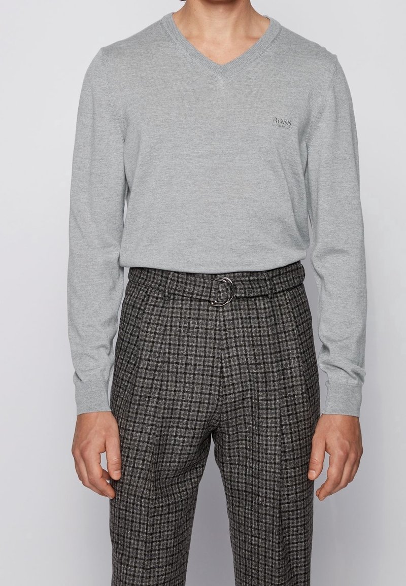 Grauer V-Ausschnitt-Pullover aus weichem Stoff, kombiniert mit karierten, hochtaillierten Hosen, die einen Gürtel mit einer runden Schnalle haben.