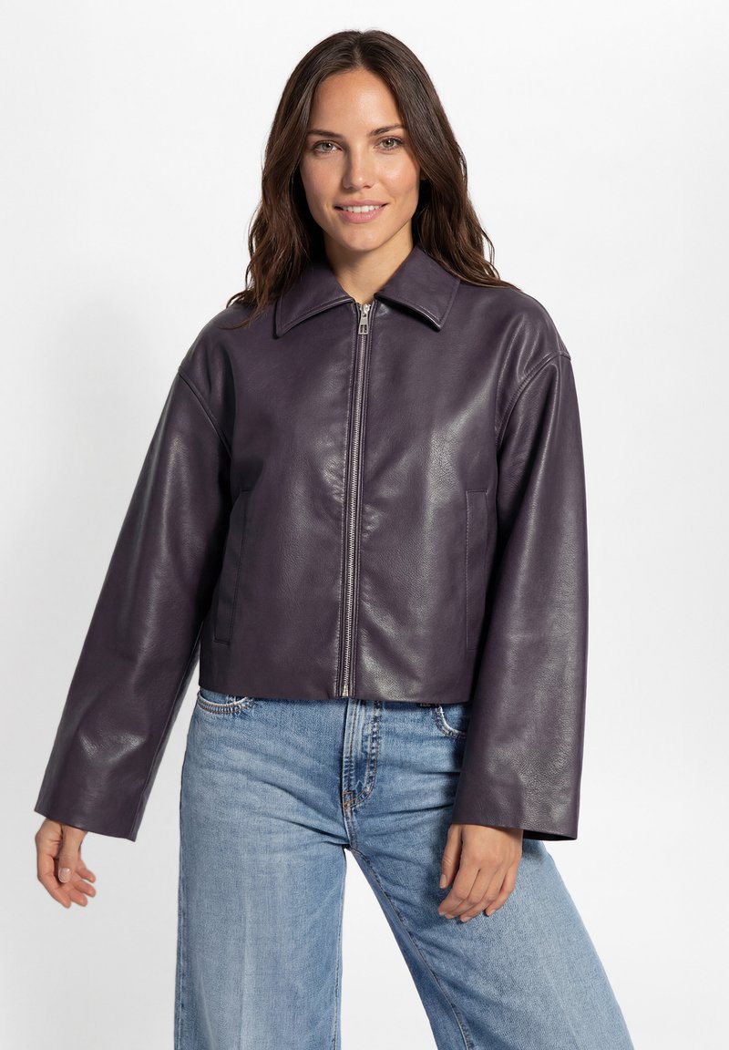 Vrouw met lang bruin haar, gekleed in een donkerpaarse leren jas met ritssluiting en lichtblauwe spijkerbroek, glimlachend tegen een witte achtergrond.