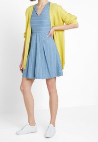 Light blue striped dress with a flared skirt, paired with a textured yellow cardigan and white sneakers. Smooth fabric and casual design.