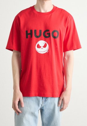 Mies, jolla on punainen T-paita, jossa lukee "HUGO" ja valkoinen pääkallokuvio, yhdistettynä vaaleansinisiin farkkuihin, kuvattuna kaulasta lanteisiin.