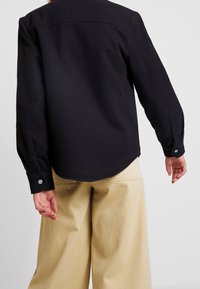 Czarna koszula z długim rękawem, o zaokrąglonym dołem i zapinanych mankietach, zestawiona z beżowymi spodniami o szerokich nogawkach. Prosty i funkcjonalny design.