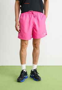 Bañador rosa con cinturilla elástica y cordón, que presenta bolsillos laterales y un pequeño logo blanco de Nike. Combinado con zapatos negros.