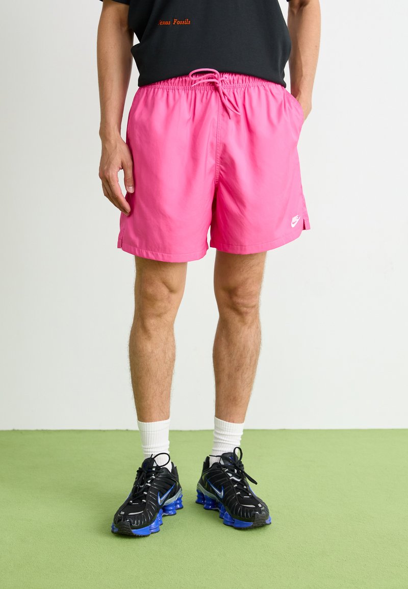 Bañador rosa con cinturilla elástica y cordón, que presenta bolsillos laterales y un pequeño logo blanco de Nike. Combinado con zapatos negros.