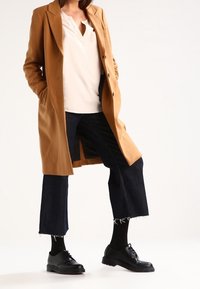 Cappotto beige sartoriale con design a un petto, abbinato a un top bianco e jeans neri tagliati. Scarpa nera stringata completa l'outfit.