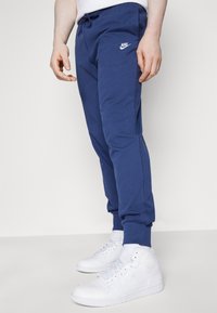 Tmavě modré tepláky značky Nike s elastickými manžetami, stahovací šňůrkou v pase a bílým logem, doplněné bílými kotníkovými teniskami.