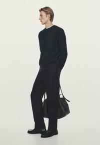 Maglione nero a coste, pantaloni scuri, scarpe nere e una grande borsa texture. Figura singola contro uno sfondo semplice.