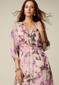 Mujer con pelo largo y castaño lleva un vestido cruzado con estampado floral en tonos pastel, con mangas abullonadas, y sostiene su cuello, complementado con pendientes y una pulsera de cuentas.