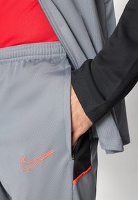 Nike treningsklær i lysegrått stoff, med jakke med glidelås, i røde og svarte detaljer, samt en oransje Nike-logo på lommen.