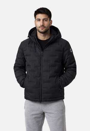 Uomo che indossa una giacca imbottita nera con cappuccio e zip e pantaloni della tuta grigio chiaro, in piedi con le mani nelle tasche della giacca.