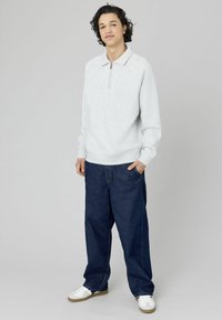 Jeune homme aux cheveux bouclés portant un sweat-shirt gris clair à fermeture éclair quart, un pantalon en denim noir ample et des baskets blanches, se tenant contre un fond gris.