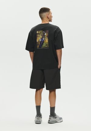 Mężczyzna stoi, ubrany w czarny, oversize'owy T-shirt z kolorowym nadrukiem natury i napisem na plecach, czarne szorty, szare skarpetki i szare trampki.