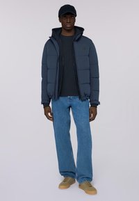 Giacca puffer blu navy con tasche con zip sopra un felpa scura, abbinata a jeans azzurri e sneaker beige. Il modello indossa un cappellino coordinato.