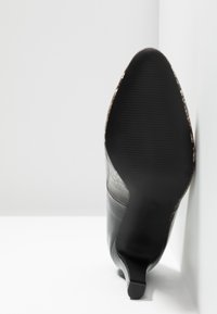 Bottine en cuir noir pointue avec un talon bas, semelle tournée vers l'avant, posée contre un angle blanc entre un mur et le sol.