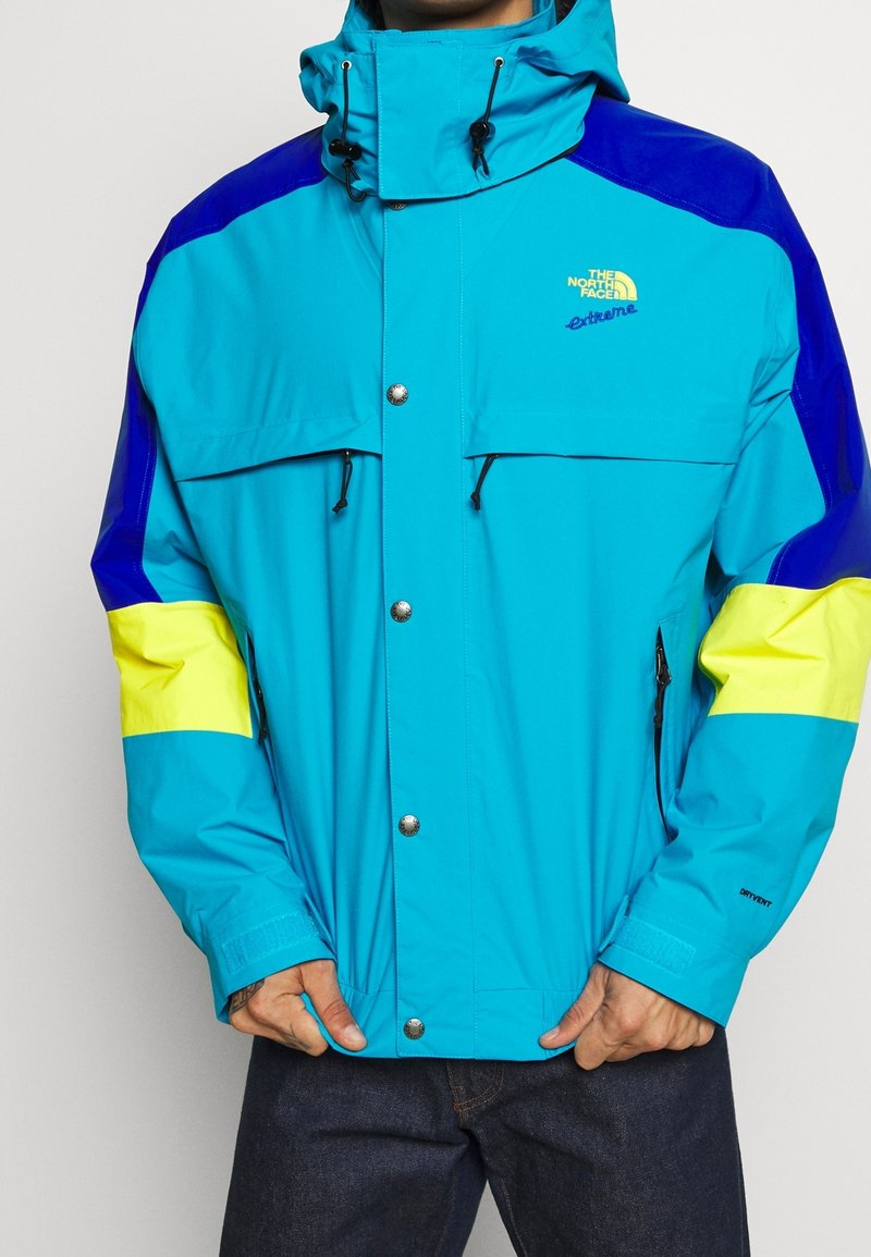 Jasnoniebieska turkusowa kurtka z niebieskimi i żółtymi akcentami, wyposażona w wysoki kołnierz, zatrzaski, regulowane sznurki oraz zapinane na zamek kieszenie.