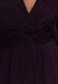 Głęboki fioletowy strój z teksturowanej tkaniny, z dekoltem w kształcie litery V i przewiązaną wstążką w talii. Swobodnie opadające plisowania dodają ruchu.