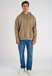Sudadera marrón con bolsillo frontal, capucha con cordón y logo. Combinada con jeans rectos azules y zapatillas blancas sobre un fondo liso.