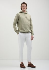 Felpa verde chiaro con tasca anteriore, abbinata a pantaloni bianchi a sigaretta e scarpe marroni con lacci, su uno sfondo neutro.