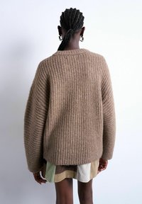 Personne aux cheveux tressés portant des boucles d'oreilles anneaux, un pull marron côtelé surdimensionné et une jupe courte à blocs de couleurs, vue de dos sur un fond clair.