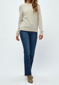 Kvinna klädd i en beige tröja, raka blå jeans och bruna ankelstövlar, stående mot en enkel ljus bakgrund.