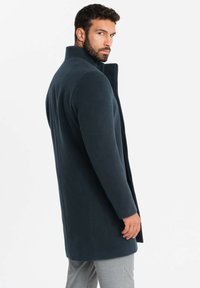 Cappotto blu navy realizzato in tessuto morbido, con un colletto alto, taglio dritto e tasche laterali, mostrato da dietro con pantaloni grigi.