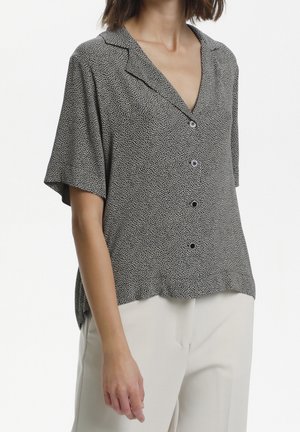 Kobieta w krótkim rękawie, czarno-białej koszuli w kropki zapinanej na guziki, w beżowych spodniach, stojąca na białym tle.