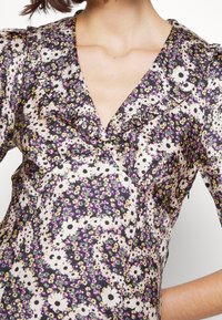 Blouse fleurie avec un col à volants, fond noir orné de fleurs blanches et violettes, tissu doux et silhouette ajustée.