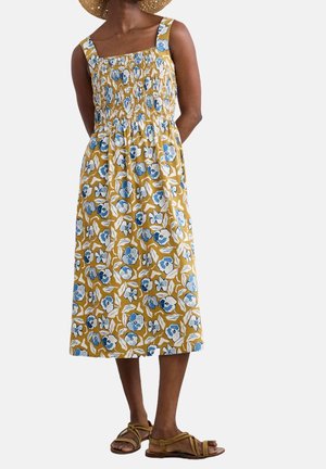 Femme portant une robe midi sans manches jaune moutarde avec un imprimé floral bleu et blanc, des sandales marron et un chapeau de paille à large bord.