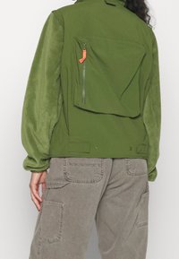 Olivově zelená bunda se měkkými, texturovanými rukávy. Obsahuje velkou zadní kapsu a kapsu na hrudi se zipem a oranžovým táhlem. Nositelská postava je v šedých cargo kalhotách.